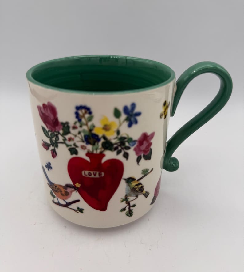 May include: A ceramic mug with a green handle and rim. The mug features a red heart-shaped vase filled with flowers and the word "LOVE". Colorful birds and floral accents decorate the off-white surface.