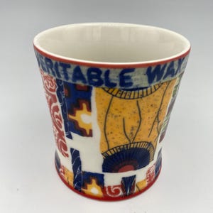 K&ouml;nnte beinhalten: Eine wei&szlig;e Keramiktasse mit einem geometrischen Muster in Rot, Blau und Gelb. Die Tasse tr&auml;gt den Schriftzug "VERITABLE WAX" auf der Seite.