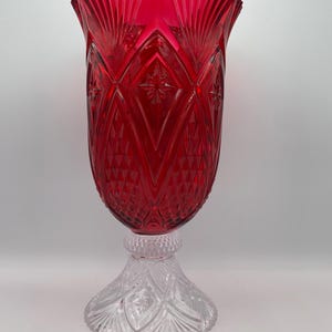 May include: A ruby red glass candle holder with a clear, ornate base. The red glass has a diamond and starburst pattern. The clear base has a detailed, cut glass design. The candle holder is a decorative home accent.