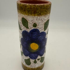 May include: A cylindrical ceramic vase with a textured white background, adorned with a large blue flower with a yellow center and green leaves. The rim is brown, and the base is a darker green.