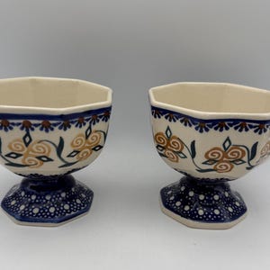 May include: Two ceramic pedestal bowls. The bowls have a cream-colored interior and are decorated with blue, brown, and green floral patterns. The bases are dark blue with white polka dots. The bowls are approximately 4 inches tall.