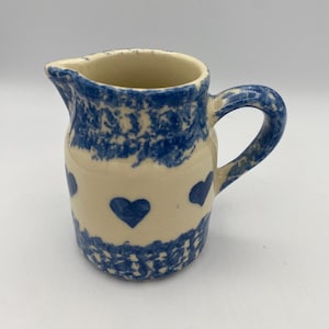 May include: Cream-colored ceramic pitcher with blue heart designs. The pitcher has a blue handle and a blue rim. The base of the pitcher has a blue patterned design. The pitcher is a decorative kitchenware item.