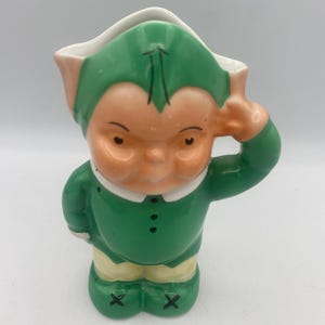 May include: A vintage ceramic figurine of a leprechaun. The figurine is primarily green with a peach-colored face and hands. It is wearing a green hat and jacket with a white collar and yellow pants. The leprechaun is posed with one hand on its head.