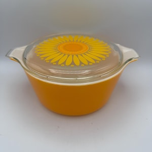 May include: An orange Pyrex bowl with a clear glass lid featuring a yellow sunflower design. The bowl has two handles and is set against a white background. A vintage kitchenware item.