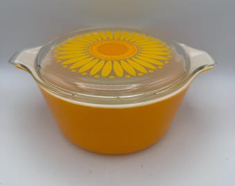 May include: An orange Pyrex bowl with a clear glass lid featuring a yellow sunflower design. The bowl has two handles and is set against a white background. A vintage kitchenware item.