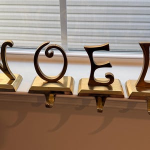May include: A set of four gold-colored metal stocking holders shaped as the letters N, O, E, and L. Each letter is mounted on a rectangular base with a downward-facing hook extending from the front edge for hanging stockings.