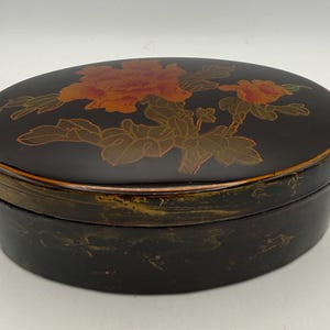 May include: An oval black lacquer box featuring a hand-painted floral design on the lid in shades of orange, red, and gold. The sides of the box display a distressed gold-toned finish with visible brush strokes and a smooth, glossy surface.