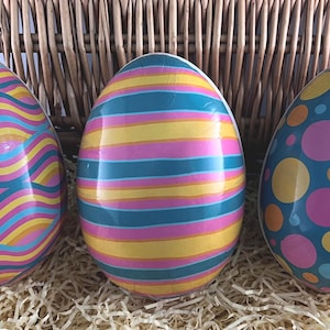 May include: Three patterned Easter eggs in a wicker basket. One egg has wavy lines in pink, yellow, and blue. Another has horizontal stripes in yellow, pink, and teal. The third egg is teal with pink, yellow, and orange polka dots.