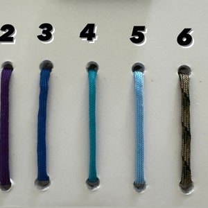 May include: A display of seven cords in various colors. The cords are black, purple, blue, teal, light blue, camouflage, and tan. Each cord is approximately 4 inches long and has a dark gray cap at the bottom.