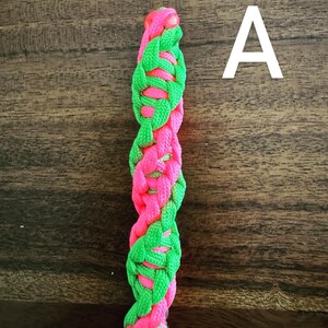 May include: A braided cord in a vertical orientation, featuring alternating sections of bright pink and neon green. The letter "A" is visible in the upper right corner. The cord appears to be made of a woven material.