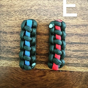 May include: Two braided paracord keychains. One is black with a blue stripe, the other is black with a red stripe. The keychains are on a wooden surface.