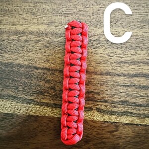 May include: A red and black woven paracord keychain fob. The fob is rectangular and features a tight weave pattern. The letter "C" is in white in the background.
