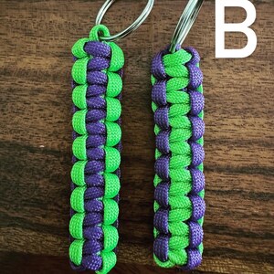 May include: Two handmade keychains with a woven design. The keychains are made of green and purple paracord and feature a silver key ring. The letter "B" is visible in the background.