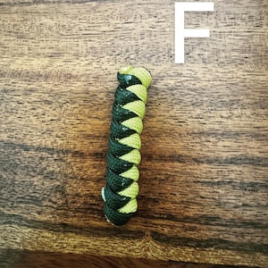 May include: A braided cord in black and yellow-green, with the letter "F" in white in the background. The cord is tightly woven, creating a textured pattern. The background is a wooden surface.