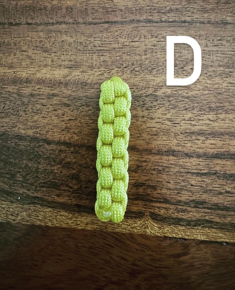 May include: A lime green, woven paracord keychain fob. The fob is rectangular and features a tight, basket-weave pattern. The background is a dark, wooden surface with visible grain.