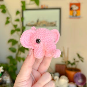 May include: A small, handmade pink crochet elephant with large ears and black eyes. The plush toy is held in a hand, showcasing its soft texture and detailed craftsmanship. The elephant is a charming, whimsical creation.