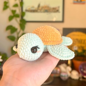 May include: A handmade crochet turtle with a light blue head and flippers, and an orange shell. The turtle has large black eyes and small pink cheeks. The plush toy is designed to look like a cartoon turtle.