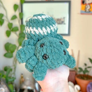 Puede incluir: Un peluche de pulpo tejido en color turquesa con una cabeza a rayas blancas y turquesas. El peluche tiene ojos de botón negros y un aspecto suave y texturizado. Este artículo hecho a mano es un juguete lindo y tierno.