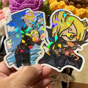 Op de afbeelding: Twee holografische stickers met anime-personages. De eerste sticker toont een blond mannelijk personage met een zwart jack en een blauwe achtergrond. De tweede sticker toont een blond vrouwelijk personage met een zwart jack en een zwarte achtergrond.