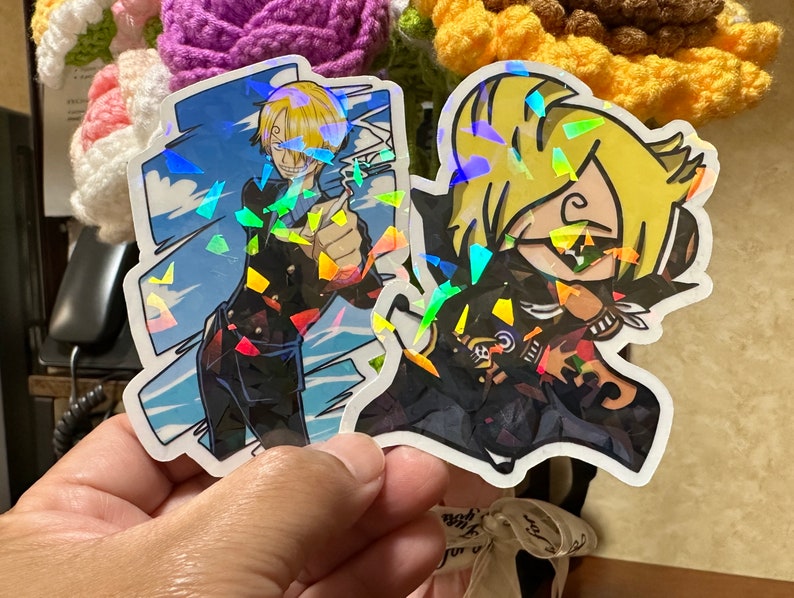 Op de afbeelding: Twee holografische stickers met anime-personages. De eerste sticker toont een blond mannelijk personage met een zwart jack en een blauwe achtergrond. De tweede sticker toont een blond vrouwelijk personage met een zwart jack en een zwarte achtergrond.