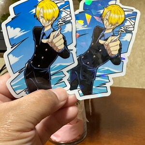 Op de afbeelding: Twee kleurrijke stickers met een anime-personage met blond haar, een zwart pak en een blauwe stropdas. Het personage wijst met zijn rechterhand. De stickers hebben een holografisch effect.