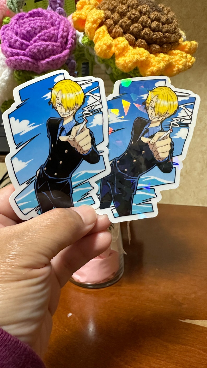 Op de afbeelding: Twee kleurrijke stickers met een anime-personage met blond haar, een zwart pak en een blauwe stropdas. Het personage wijst met zijn rechterhand. De stickers hebben een holografisch effect.