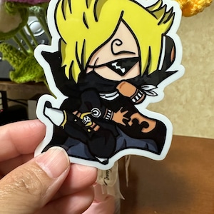 Op de afbeelding: Een sticker met een cartoonfiguur met geel haar, die een zwart masker en een zwart en bruin pak draagt. Het figuur houdt een zwaard vast en heeft een schedel en gekruiste beenderen op zijn arm.