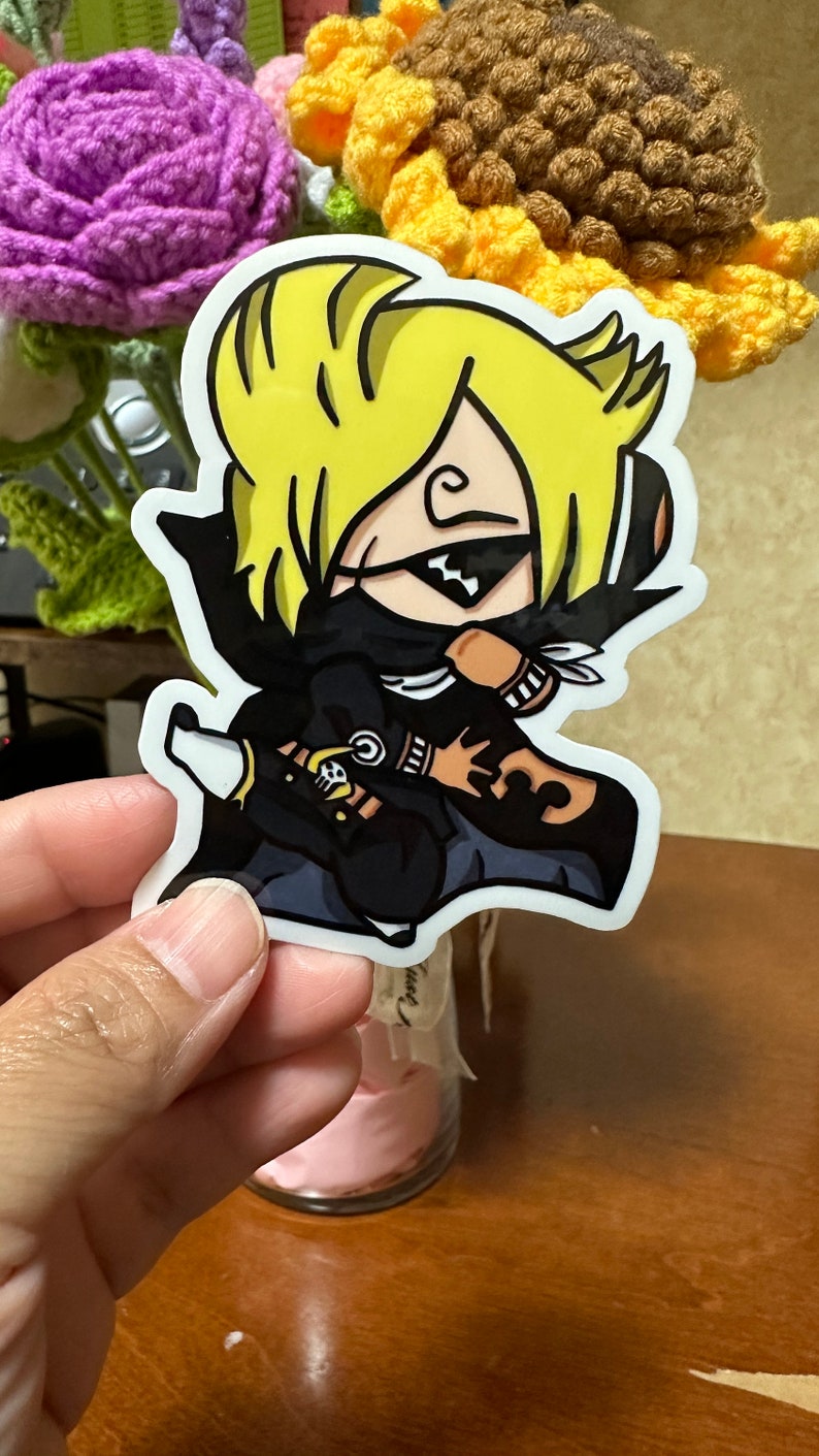 Op de afbeelding: Een sticker met een cartoonfiguur met geel haar, die een zwart masker en een zwart en bruin pak draagt. Het figuur houdt een zwaard vast en heeft een schedel en gekruiste beenderen op zijn arm.