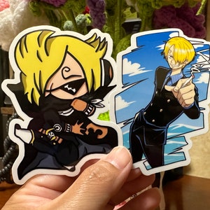 Op de afbeelding: Twee anime-personage stickers. E&eacute;n sticker toont een personage met geel haar, een zwart masker en een zwart-witte outfit. De andere sticker toont een personage met geel haar, een blauw shirt en een zwart jack. De achtergrond van de sticker is blauw met witte wolken.