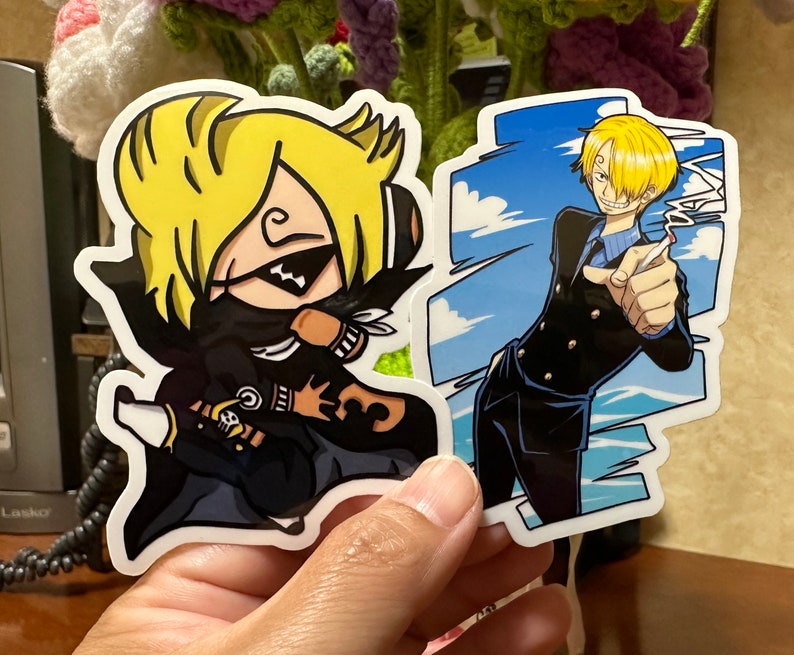 Op de afbeelding: Twee anime-personage stickers. E&eacute;n sticker toont een personage met geel haar, een zwart masker en een zwart-witte outfit. De andere sticker toont een personage met geel haar, een blauw shirt en een zwart jack. De achtergrond van de sticker is blauw met witte wolken.