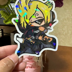 Op de afbeelding: Een holografische sticker met een cartoonfiguur met blond haar, gekleed in een zwart-witte outfit met een schedel- en kruisbeenderontwerp. Het personage draagt ​​een zwart masker met een witte streep over de ogen.