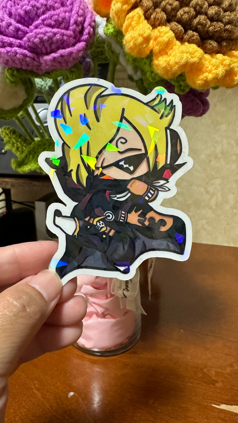 Op de afbeelding: Een holografische sticker met een cartoonfiguur met blond haar, gekleed in een zwart-witte outfit met een schedel- en kruisbeenderontwerp. Het personage draagt ​​een zwart masker met een witte streep over de ogen.