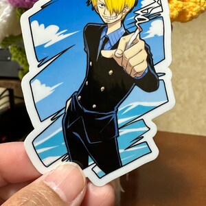 Op de afbeelding: Een sticker met een cartoonfiguur met blond haar, die een zwart pak met gouden knopen, een blauw overhemd en een blauwe das draagt. Het personage houdt een sigaret in zijn rechterhand en heeft een ondeugende uitdrukking op zijn gezicht. De achtergrond is een blauwe lucht met witte wolken.