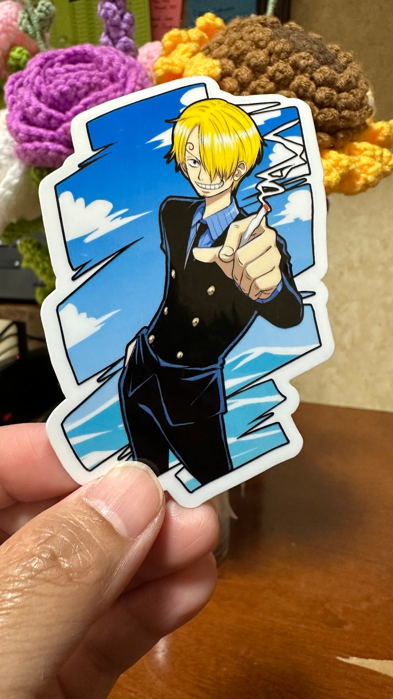 Op de afbeelding: Een sticker met een cartoonfiguur met blond haar, die een zwart pak met gouden knopen, een blauw overhemd en een blauwe das draagt. Het personage houdt een sigaret in zijn rechterhand en heeft een ondeugende uitdrukking op zijn gezicht. De achtergrond is een blauwe lucht met witte wolken.