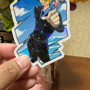Op de afbeelding: Een holografische sticker met een cartoonfiguur met blond haar, die een zwart pak en een blauw shirt draagt en een sigaret vasthoudt. Het personage staat voor een blauwe lucht met witte wolken.
