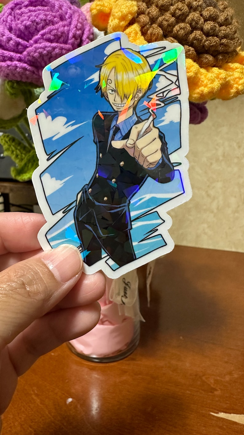 Op de afbeelding: Een holografische sticker met een cartoonfiguur met blond haar, die een zwart pak en een blauw shirt draagt en een sigaret vasthoudt. Het personage staat voor een blauwe lucht met witte wolken.