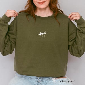 May include: Olive green sweatshirt with a white embroidered ant design. The sweatshirt is a crew neck style and has long sleeves. The text "military green" is at the bottom of the image.