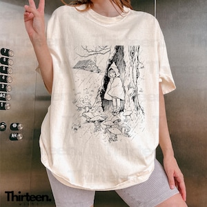 May include: A cream-colored t-shirt with a black and white illustration of a girl standing under a tree in the rain. The illustration includes a small house and mushrooms. The text "Thirteen. wishes" is printed on the bottom left corner of the shirt.
