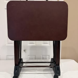 May include: A rectangular dark brown folding TV tray table with a smooth surface and a black metal handle on top. The table is supported by four thin dark brown metal legs connected to a black plastic base featuring four small rolling casters.