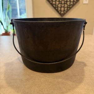 May include: A black cast iron pot with a wide, circular opening and a thick, rounded base. The pot features a sturdy, thin metal handle attached to two small side lugs, allowing it to swing freely. The exterior surface has a matte, textured finish.