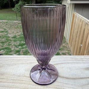 May include: A tall, translucent purple glass goblet featuring a vertical ribbed pattern along the bowl. The glass has a wide, circular base with a star-shaped design at the center where the stem connects to the foot, creating a clear, elegant profile.