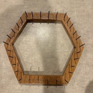 May include: A hexagonal wooden frame featuring a series of evenly spaced metal nails protruding from the top surface. The wood has a natural brown grain finish, and the metal nails are arranged along the perimeter of the six-sided structure.