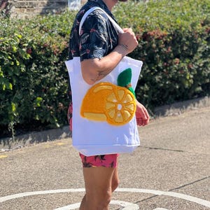 Op de afbeelding: Witte draagtas met een geborduurd sinaasappelschijf-ontwerp. De sinaasappelschijf is geel en oranje met een groen blad. De tas heeft witte bandjes. De persoon draagt een shirt met patroon en een roze korte broek.
