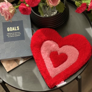 May include: A red heart-shaped pillow with a pink heart inside. The pillow is made of a soft, fuzzy material. The pillow is sitting on a black table with a book that says "Goals" on it.