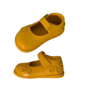 Puede incluir: Zapatos Mary Jane de color amarillo mostaza con cierre de correa. Los zapatos tienen un diseño floral en la puntera y el lateral. Estos zapatos son para niños pequeños.