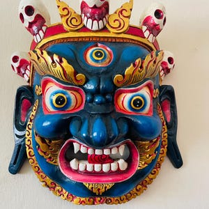 Puede incluir: Una máscara de madera tallada a mano con una expresión feroz. La máscara presenta una cara azul con detalles dorados, ojos grandes y una amplia sonrisa con dientes. Adornando la parte superior hay varios adornos en forma de calavera con detalles rojos y blancos. Una llamativa pieza de decoración mural.