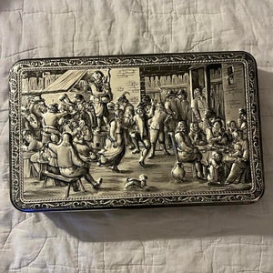 May include: A silver-plated metal box with a raised image of a group of people gathered in a tavern. The image is in black and white and features a detailed depiction of the people and their surroundings.