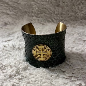 May include: A wide, black leather cuff bracelet with a gold-toned, circular design featuring a cross in the center. The bracelet has a textured surface.