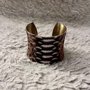 May include: A wide, brown snakeskin cuff bracelet with a brass base. The bracelet is made of real snakeskin and has a unique, textured design.