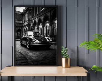 May include: Black and white photograph of a classic black Porsche car parked on a cobbled street in front of an old building.